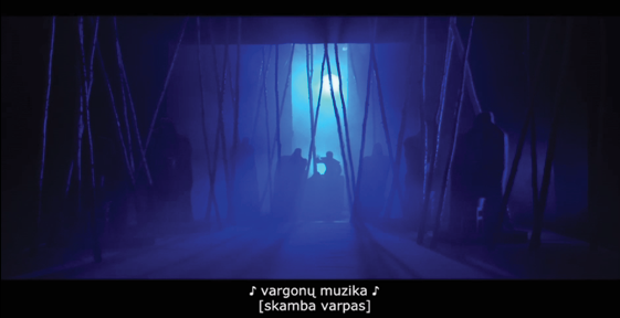 In a dark stage setting with deep blue lighting, multiple tall, rope-like cords hang from the ceiling in rows. Silhouettes of actors stand among these ropes. Subtitles in Lithuanian at the bottom of the image read “vargonų muzika” (organ music) and “skamba varpas” (a bell rings).
