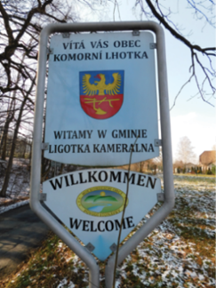 Dwa zdjęcia obok siebie. Na pierwszym zdjęciu jest krajobraz z drogą, skrzyżowaniem o ruchu okrężnym oraz ekranami akustycznymi drogi szybkiego ruchu w tle. Na pierwszy plan wysuwają się dwie tablice drogowe „koniec gminy Bystřice” w języku czeskim oraz „koniec gminy Bystrzyca“ w języku polskim. Czeską tablicę zainstalowano na metalowym słupku wyżej od polskiej. Wielkością, kształtem, kolorami i czcionką są obydwie identyczne. Na drugim zdjęciu zobaczyć można „tablicę powitalną“, zainstalowaną przy drodze w otoczeniu drzew. W środku metalowej tablicy w kształcie prostokątu i niewielkiego trójkątu widnieje znak gminy Ligotka Kameralna (Komorní Lhotka), wokół którego zamieszczono napisy w języku czeskim, polskim, niemieckim i angielskim „Vítá vás obec Komorní Lhotka / Witamy w gminie Ligotka Kameralna / Willkommen / Welcome.