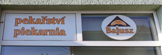Zdjęcie przedstawia fragment witryny sklepowej. W prawej części jest nazwisko/nazwa sklepu „Bajusz” i logo rogalika (pieczywo). Wlewo są dwa napisy – u góry czeski „pekařství”, pod nim polski „piekarnia”. Graficznie są wszystkie napisy tożsame – jednakowej wielkości i pomarańczowego koloru.