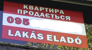 Фото 7. На окне частного дома размещена прямоугольная вывеска красно-белого цвета о продаже квартиры. В верхней части белыми буквами на красном фоне написано «КВАРТИРА ПРОДАЄТЬСЯ» на украинском. По центру красным цветом на белом фоне указан номер телефона украинского оператора связи, начинающийся с кода «095» (последующие цифры скрыты автором этой статьи для сохранения конфиденциальности). В нижней части на красном фоне белыми буквами написано “LAKÁS ELADÓ” на венгерском. В этом объявлении нет грамматических ошибок.