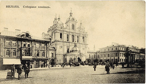 A historic sepia-toned street scene shows a large ornate church with a dome and towers in the center, flanked by multi-story buildings, with people walking and standing on a cobblestone square.
Description generated by AI
