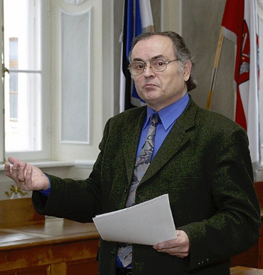 A person wearing glasses, a dark blazer, blue shirt, and patterned tie is holding papers and gesturing with one hand in a formal room with flags and large windows.
Description generated by AI