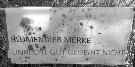 A weathered sign with spots and stains reads "BLUMENDIEB MERKE UNRECHT GUT GEDEIHT NICHT" in German.

Description generated by AI